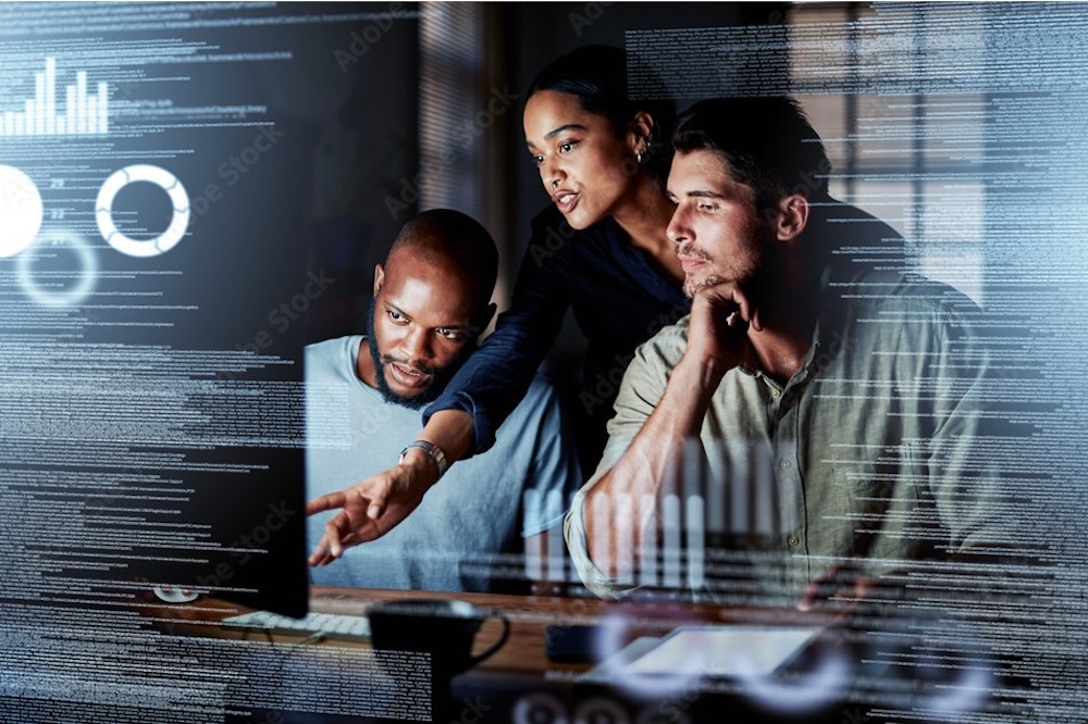 IT admins at computer Three IT admins looking at a computer with vaguely cybersecurity-esque graphics overlaid in the foreground.